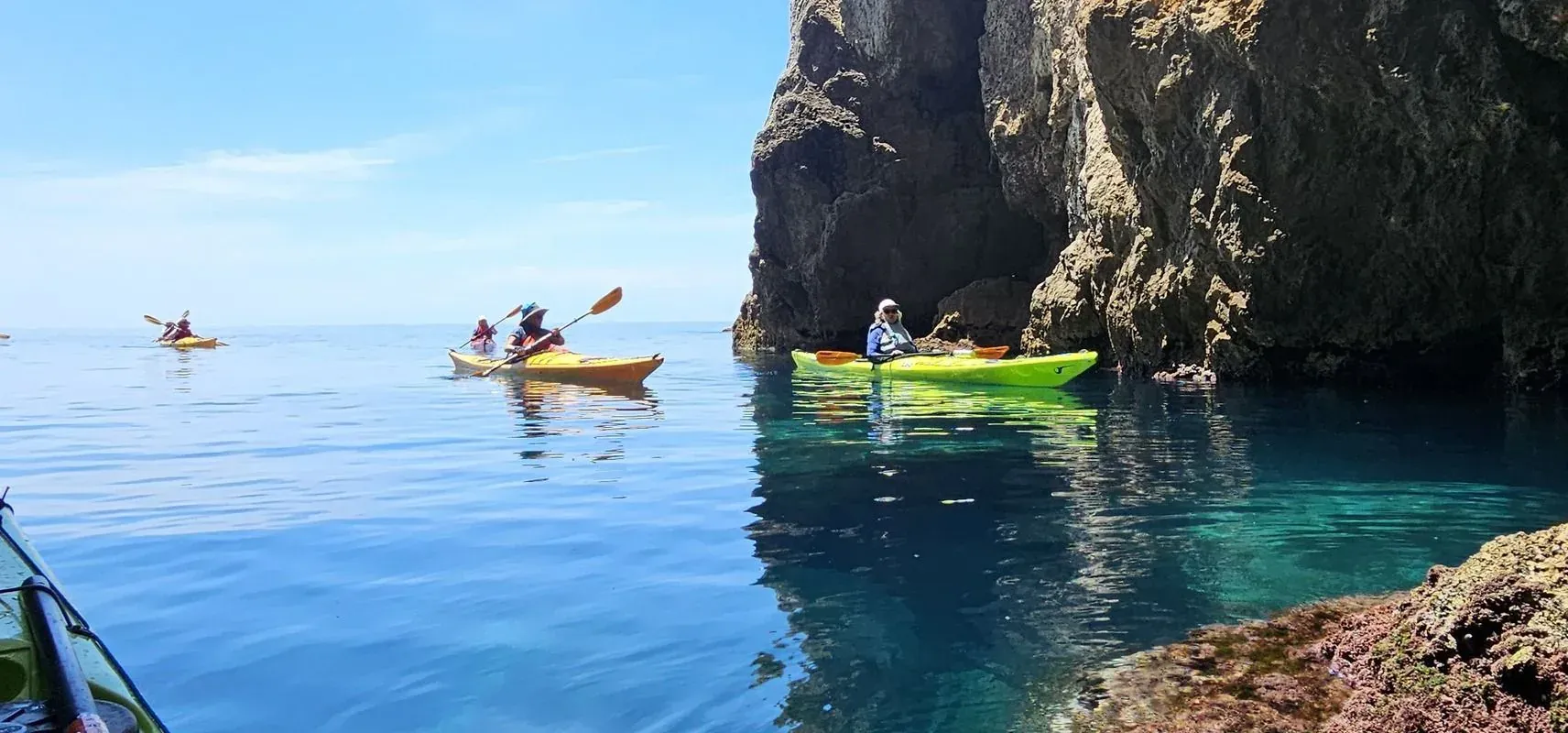 Kayakers on clear turquoise water beside a rocky cliff