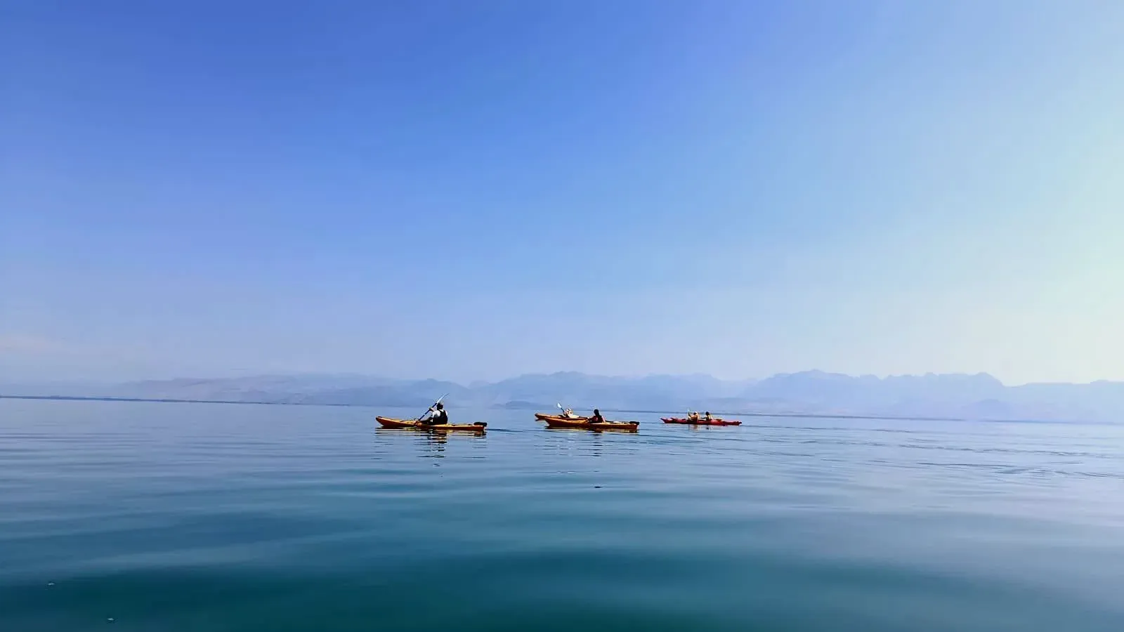 Three sea kayaks crossing glassy open water toward distant mountains