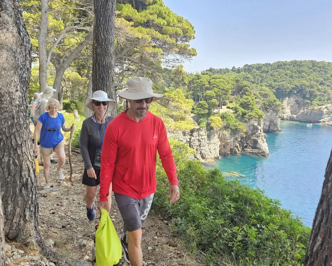 Hikers on a pine-shaded coastal trail above a turquoise cove