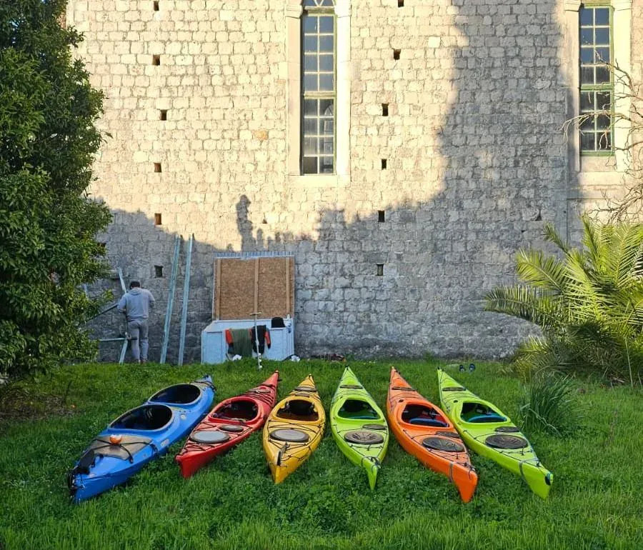 A row of colourful sea kayaks lined up on the grass beside a stone island building