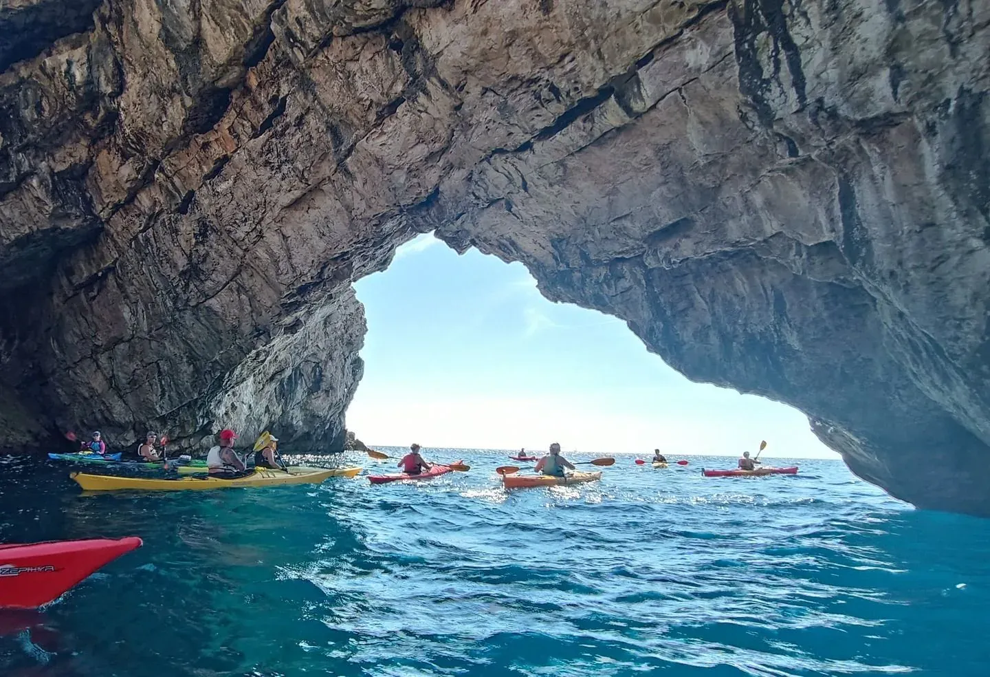 Kayakers paddling out from beneath a large sea-cave arch into open turquoise water