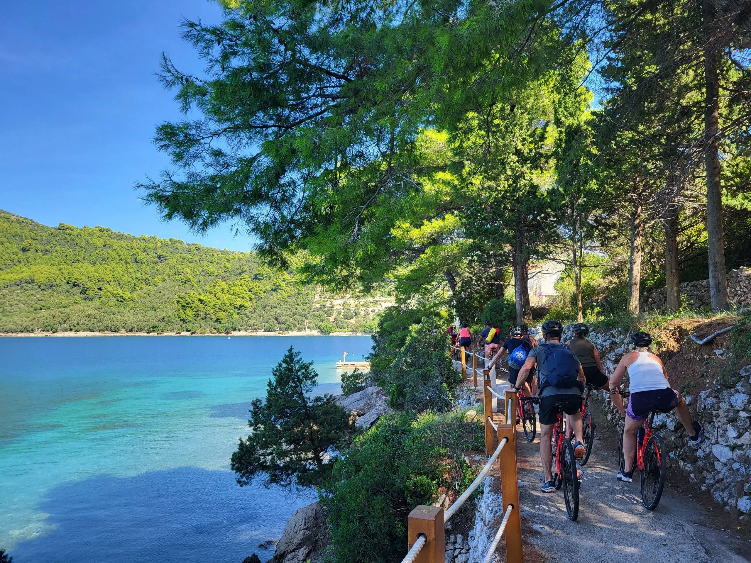 Cyclists riding a shaded coastal path above a turquoise bay