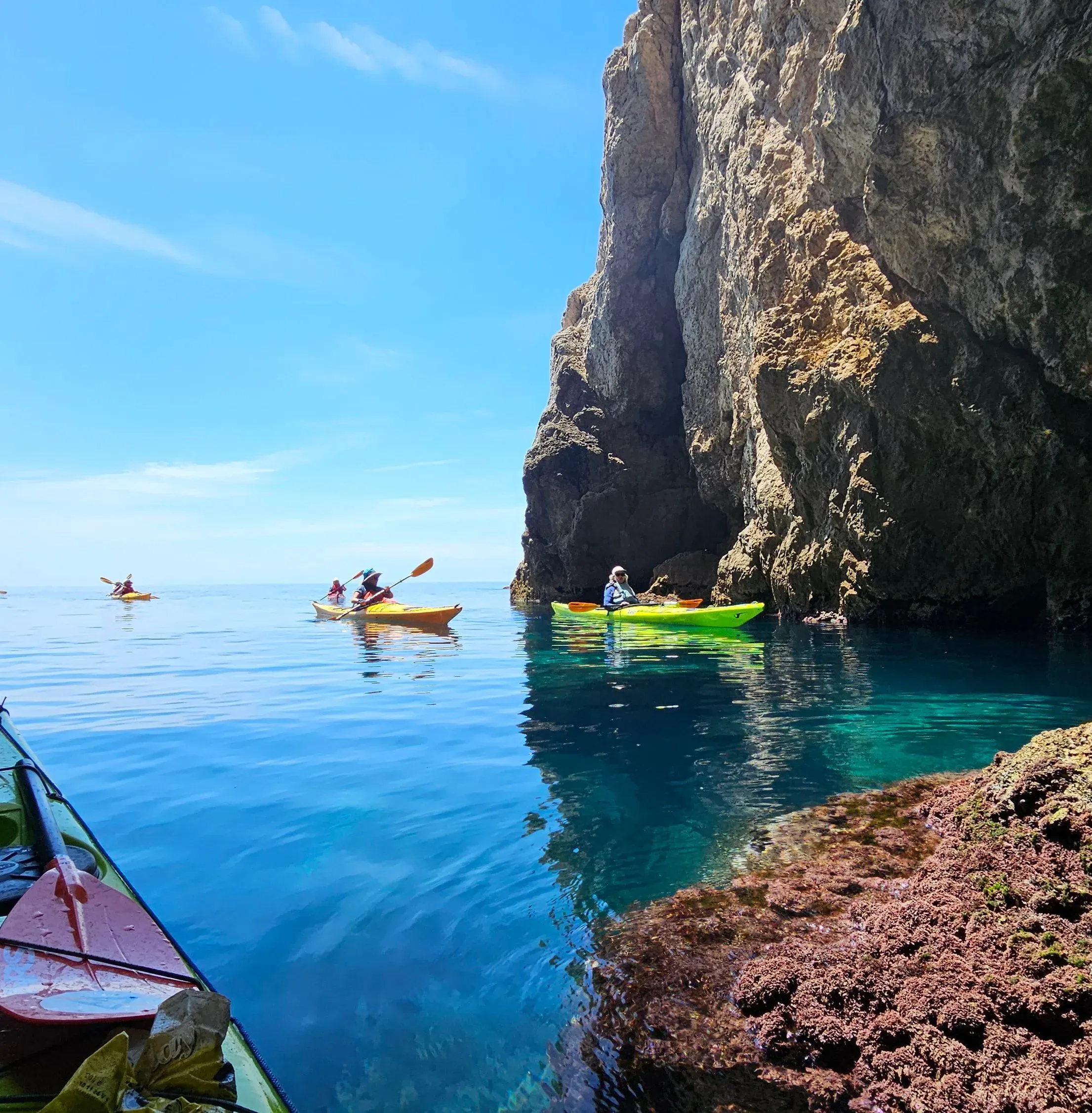 Kayakers paddling calm turquoise water below a tall coastal cliff