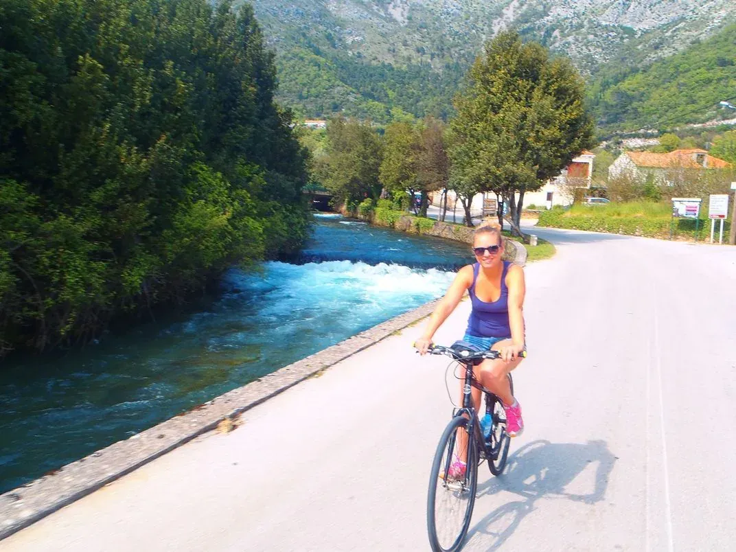 A cyclist riding a quiet road beside a fast-flowing river below bare mountains