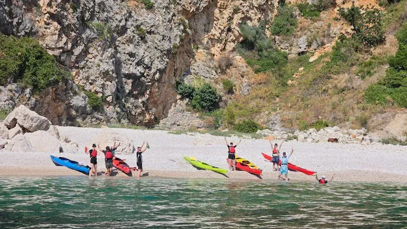 Kayakers landing on a pebble beach below a limestone cliff, paddles raised