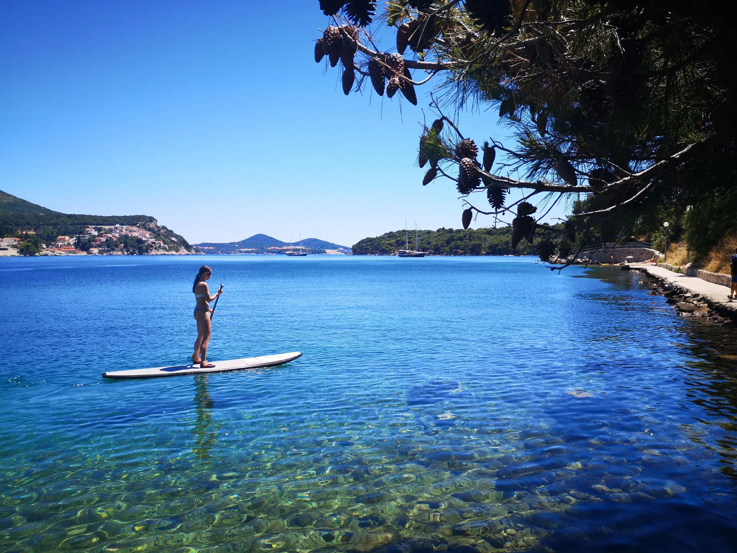 A stand-up paddleboarder crossing clear shallow water with an island town beyond