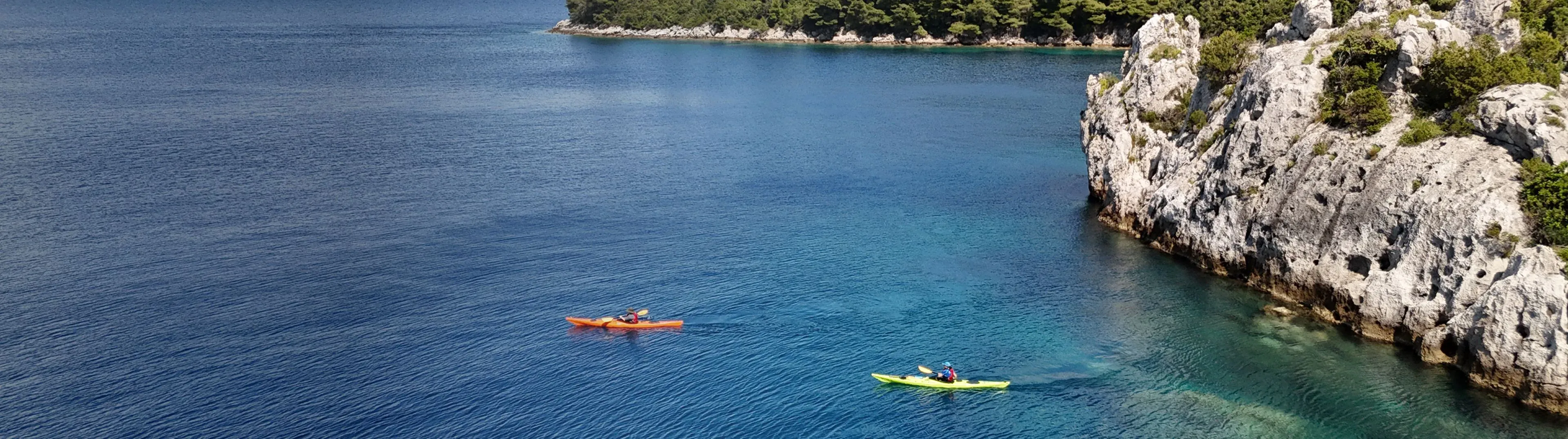 Two sea kayaks crossing deep blue water below a wooded limestone headland