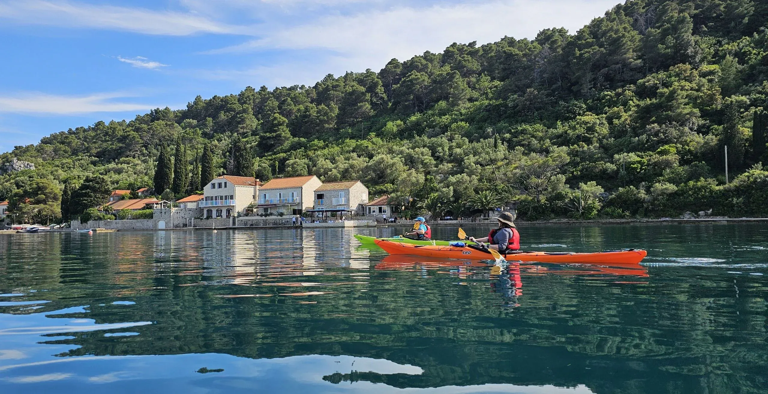 A kayaker in an orange sea kayak passing a waterfront villa below a wooded hillside