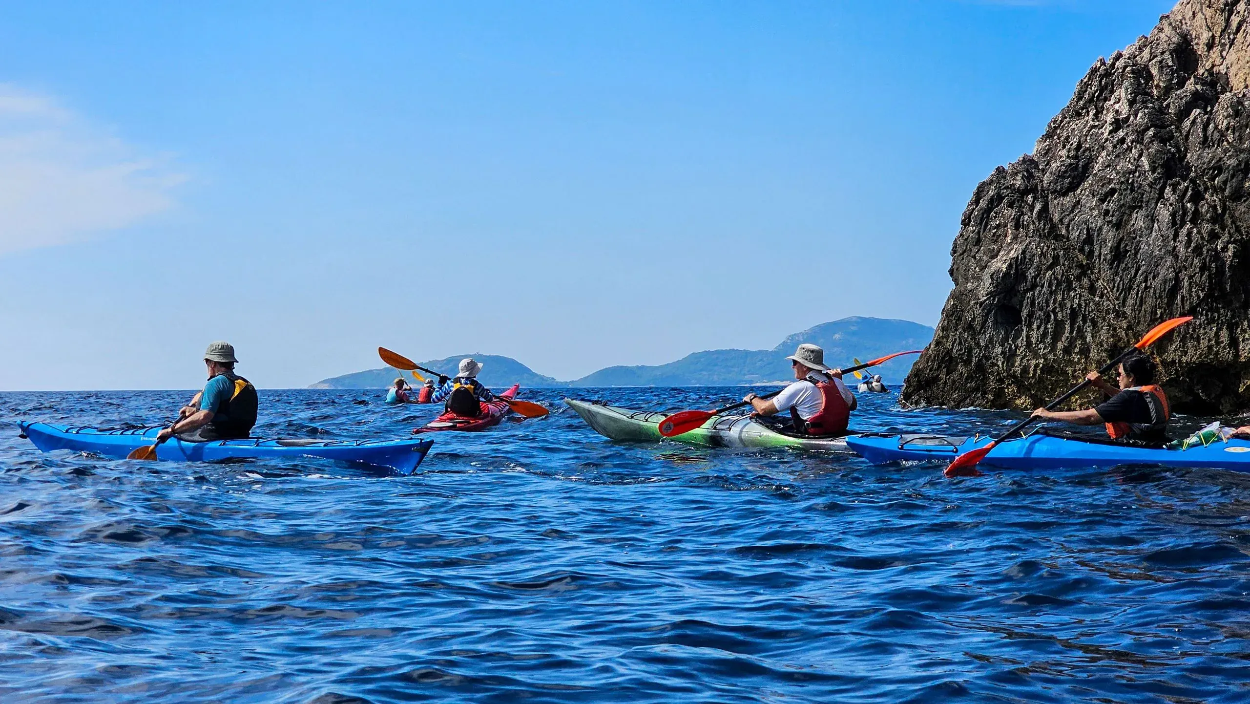 A group of sea kayakers gathered beside a rock face on open blue water