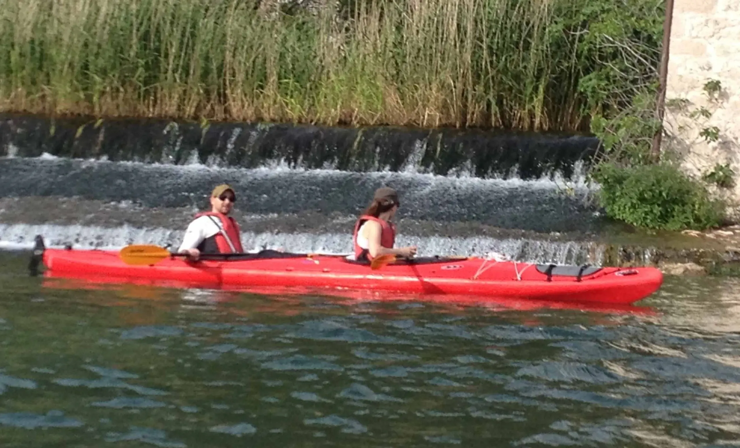 Two paddlers in a red tandem kayak on a calm river below a low weir